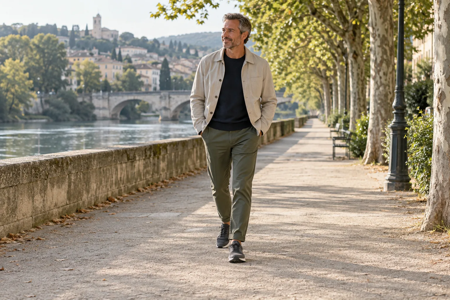 A man in olive chinos and charcoal sneakers strolling along a tree-lined riverside path